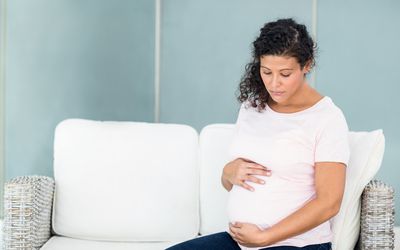 Sad pregnant woman sitting on sofa against wall